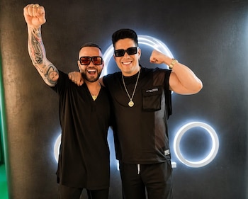 Chyno y Nacho, sonrientes, posan con gafas de sol y ropa oscura. Chyno con brazo en alto, Nacho flexionando bíceps. Fondo oscuro con anillos luminosos