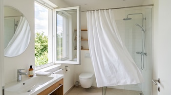 Cuarto de baño con lavabo, espejo, inodoro, ducha con cortina blanca y una ventana abierta que da al exterior.
