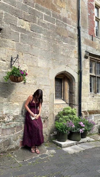 Mujer con vestido largo borgoña y sandalias, apoyada en pared de piedra. Hay un cesto de flores colgante, macetas y una ventana arqueada