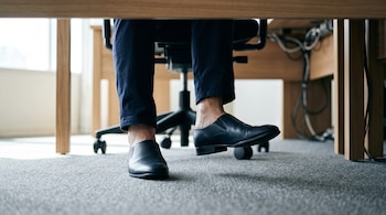 Pies y piernas de una persona con zapatos negros debajo de un escritorio, con un pie levemente elevado del suelo.