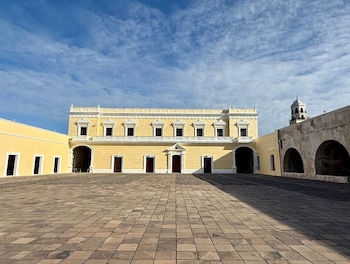Edificio colonial amarillo de dos pisos, balcón blanco, ventanas y puertas. Patio empedrado, muro con arcos y campanario bajo cielo azul