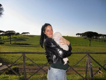 Oriana Sabatini sonríe a cámara mientras sostiene a un bebé envuelto en tela clara, al aire libre. Detrás, una valla de madera, un campo verde y árboles