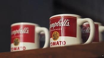 Primer plano de tazas de café blancas con la etiqueta roja y blanca de "Campbell's Condensed Tomato Soup" sobre un estante de madera oscuro