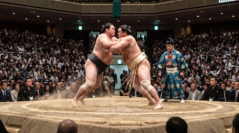 Dos luchadores de sumo en un dohyo, un árbitro de sumo, una audiencia en el fondo y un estadio interior.
