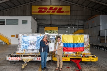 Dos mujeres sostienen un balde blanco frente a dos cargamentos de mercancía cubiertos con las banderas de Guatemala y Colombia