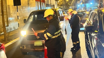 Trabajadores con chalecos reflectantes y cascos amarillos inspeccionan un vehículo oscuro en una calle iluminada de noche, con otros coches cerca
