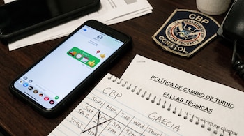 Un teléfono móvil en una mesa muestra un mensaje con un camión, reloj, calendario y pulgares arriba. Un cuaderno espiral, un parche CBP y otros papeles están junto al teléfono.