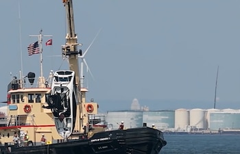 Una grúa en un barco del Cuerpo de Ingenieros del Ejército de Estados Unidos levanta una lancha blanca volcada del agua. Hay personas, una bandera estadounidense y tanques industriales