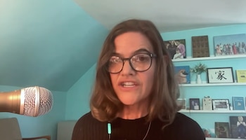 Mujer con gafas frente a un micrófono en una habitación con estanterías y una pared azul