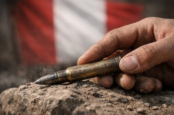 Primer plano de una mano sucia que sostiene un cartucho de fusil metálico sobre una roca polvorienta, con la bandera de Perú desenfocada al fondo.