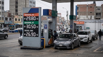 Cartel con precios de combustibles junto a surtidores de una estación de servicio con vehículos estacionados y edificios al fondo.