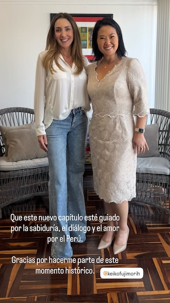 Dos mujeres de pie sonríen. Una viste blusa blanca y jeans, la otra un vestido beige de encaje. Hay sillas de mimbre y un cuadro al fondo. Texto en pantalla