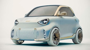 Primer plano de un prototipo de coche Smart de color gris claro con detalles dorados, faros ovalados y un diseño futurista en un fondo blanco