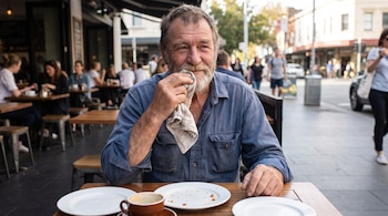 Hombre mayor sentado en una mesa de madera al aire libre, con pañuelo en la mano, tres platos blancos vacíos y una taza de café marrón.