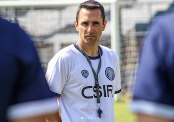 Hombre de mediana edad con camiseta blanca y ribete azul, cordón y silbato, mirando directamente. Fondo borroso de campo deportivo con red