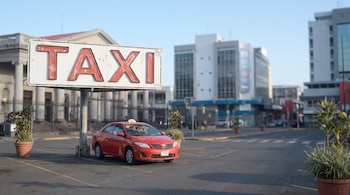 Un taxi rojo estacionado frente a un cartel grande con la palabra TAXI, en un entorno urbano con edificios y un cielo azul claro.