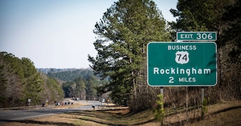 Una señal de tráfico verde con texto blanco que indica 'EXIT 306', 'BUSINESS 74', 'Rockingham' y '2 MILES'. Al fondo, una autopista con varios coches y árboles