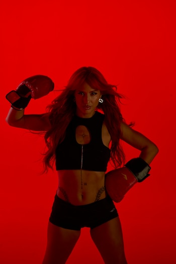 Flor Vigna posa de frente con guantes rojos de boxeo, un crop top negro y shorts, sobre un fondo rojo vibrante, con el cabello suelto
