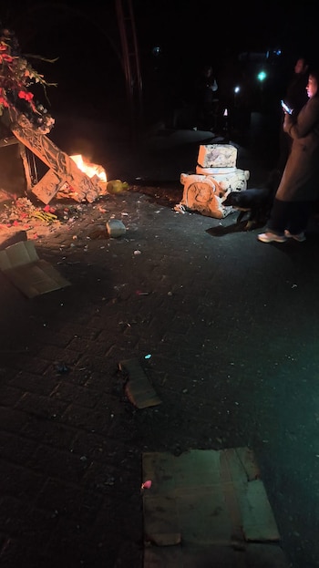 Fuego en el suelo junto a una cruz decorada con flores rojas y blancas. Hay personas, un perro, cartones y un pedestal de piedra