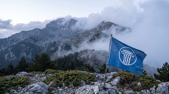 Bandera azul con logo blanco de UNESCO, montañas rocosas con nubes, vegetación baja, pinos y rocas grises.
