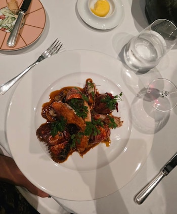 Un plato blanco de langosta en salsa sobre un mantel blanco junto a cubiertos, una copa y platos pequeños