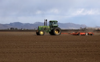 Un tractor verde John Deere tira de un equipo rojo sobre un campo de tierra marrón, con montañas y cielo nublado al fondo