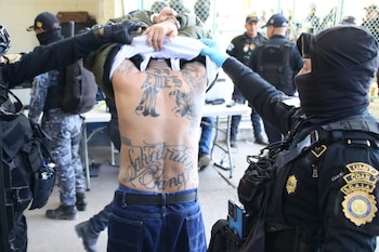 Hombres en uniforme militar y cascos, con batones, supervisan a un grupo numeroso de hombres, algunos sin camisa, en un pasillo abarrotado