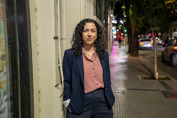 Mujer con cabello rizado, blazer azul marino y camisa rosa, de pie en acera por la noche. Al fondo, una calle con luces de vehículos y edificios