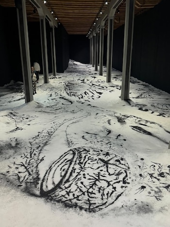 Instalación artística interior. Suelo blanco con patrones negros. Pilares grises, techo de madera y paredes oscuras. Dos personas pequeñas caminan al fondo