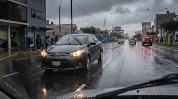 Un automóvil gris con faros encendidos sobre una avenida mojada por la lluvia, peatones con paraguas en la acera y vehículos al fondo.