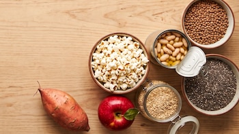 Una batata, una manzana, palomitas de maíz, frijoles en lata, lentejas, avena y semillas de chía sobre una mesa de madera