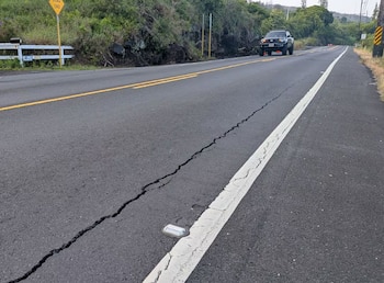 Una carretera de asfalto gris muestra una grieta longitudinal prominente. Hay líneas blancas y amarillas, un reflector vial y un camión oscuro a lo lejos