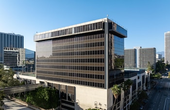 Edificio de fachada de vidrio oscuro, estructura rectangular, cielo azul despejado, otros edificios en la distancia, árboles, palmeras, calle con vehículos, puente peatonal