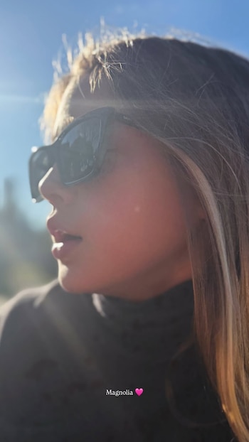 Retrato de perfil cercano de una persona con cabello castaño claro y gafas de sol oscuras, con el sol de fondo. Texto: Magnolia y un corazón rosa