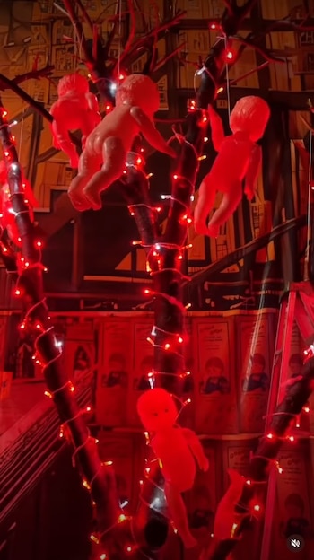 Ramas de árbol envueltas en tiras de luces rojas encendidas de las cuales cuelgan pequeñas figuras de plástico con forma de bebé