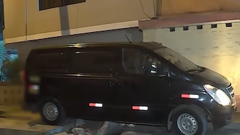 Una minivan oscura con reflectores rojos y blancos está sobre una acera rota junto a un edificio. Sus luces delanteras brillan en la oscuridad de la noche