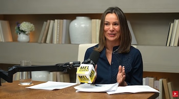 Una mujer sentada a una mesa frente a un micrófono con el logotipo de The Mel Robbins Podcast, con papeles y un vaso de agua