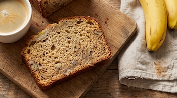Rebanada de pan de plátano con nueces, pan de plátano, taza de café, dos plátanos amarillos y un paño de tela sobre tabla de madera.
