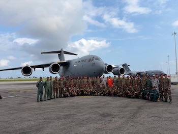 Grupo de militares uniformados posa frente a un avión de carga gris de la Fuerza Aérea de Qatar. Más aviones similares y una pista de aeropuerto bajo cielo nublado