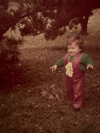 Un niño sonriente con cabello castaño claro, vestido con un enterito rojo y suéter verde, camina sobre césped. Árboles con hojas oscuras detrás