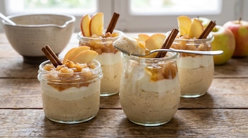Vista cenital de cuatro vasos de cristal con mousse de manzana y yogur, decorados con rodajas de manzana, canela y compota, sobre una mesa de madera.