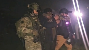 Imagen nocturna de tres hombres; dos, uno en uniforme camuflado, escoltan a un tercero con manos levantadas, visiblemente iluminados por focos brillantes
