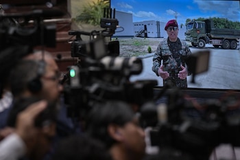 Oficial militar en uniforme, boina roja y gafas de sol en una pantalla grande, con cámaras de video y personas borrosas en primer plano