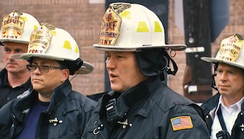 Cuatro bomberos del FDNY con cascos blancos y uniformes oscuros. Un bombero con casco de Chief en primer plano habla. Se aprecia un parche de la bandera de Estados Unidos