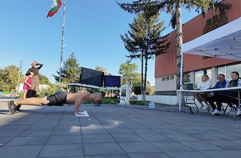 Un hombre hace flexiones en el suelo frente a dos pantallas digitales y tres personas sentadas bajo una carpa, junto a la bandera de Bulgaria