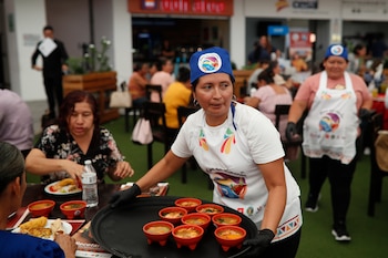 Una mujer sonriente con gorro y delantal blanco con un logo de comida, sostiene una bandeja negra con tres platos de comida. Al fondo, personas y pancartas