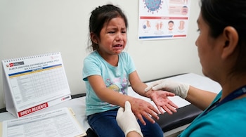 Niña llorando con erupciones rojas en manos y boca es examinada por un médico con guantes. Hay carteles informativos sobre la alerta sanitaria por Coxsackie de Perú.