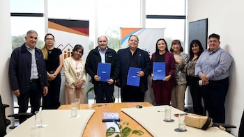Nueve personas, cinco hombres y cuatro mujeres, posan de pie en una oficina; tres de ellos en el centro sostienen carpetas azules. Una mesa de reuniones está en primer plano