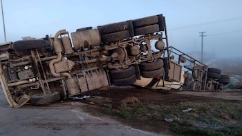 Un camión cisterna blanco yace volcado sobre el costado de una carretera, con las ruedas expuestas. El terreno es de tierra y pasto. Se ve niebla y un poste eléctrico