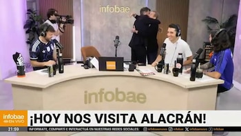 Dos hombres se abrazan en el estudio de Infobae Mundial. Varias personas están sentadas alrededor de un escritorio con micrófonos y botellas